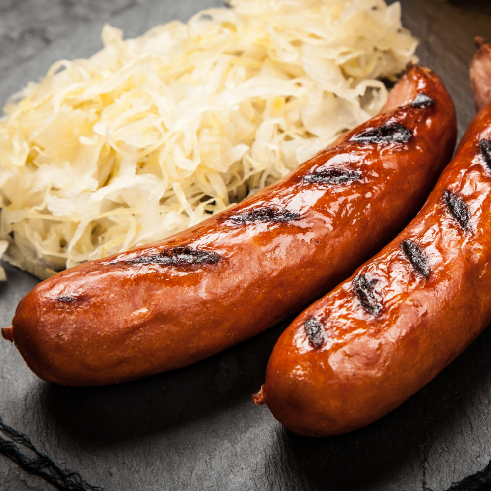 Zwei gegrillte Bratwürste mit Röstaromen, serviert auf einem dunklen Teller mit einer Portion Sauerkraut im Hintergrund.